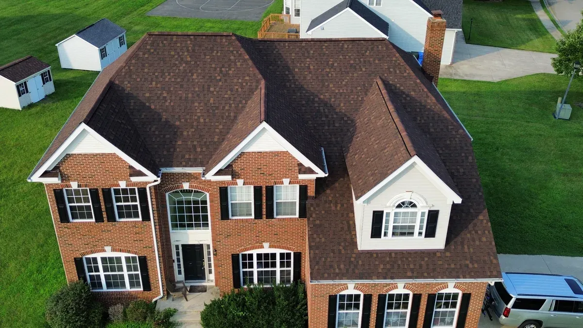 Aerial view of a brick colonial with a finished brown architectural shingle roof, two front gables, a bay dormer over the garage, and a brick chimney