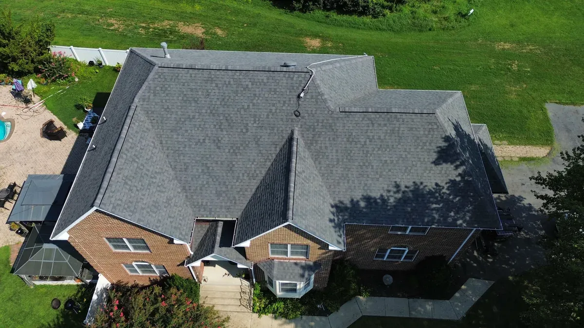 Aerial view of a large brick home with a finished grey architectural shingle roof, multiple gables and hips meeting over a center ridge, sunroom and patio at the side