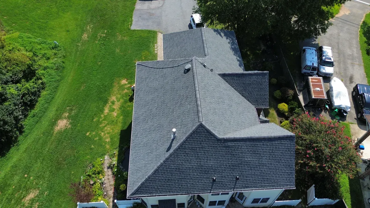 Aerial view of a finished grey architectural shingle roof on a home set against a sloped lawn, with the crew truck and dump trailer staged on the street below