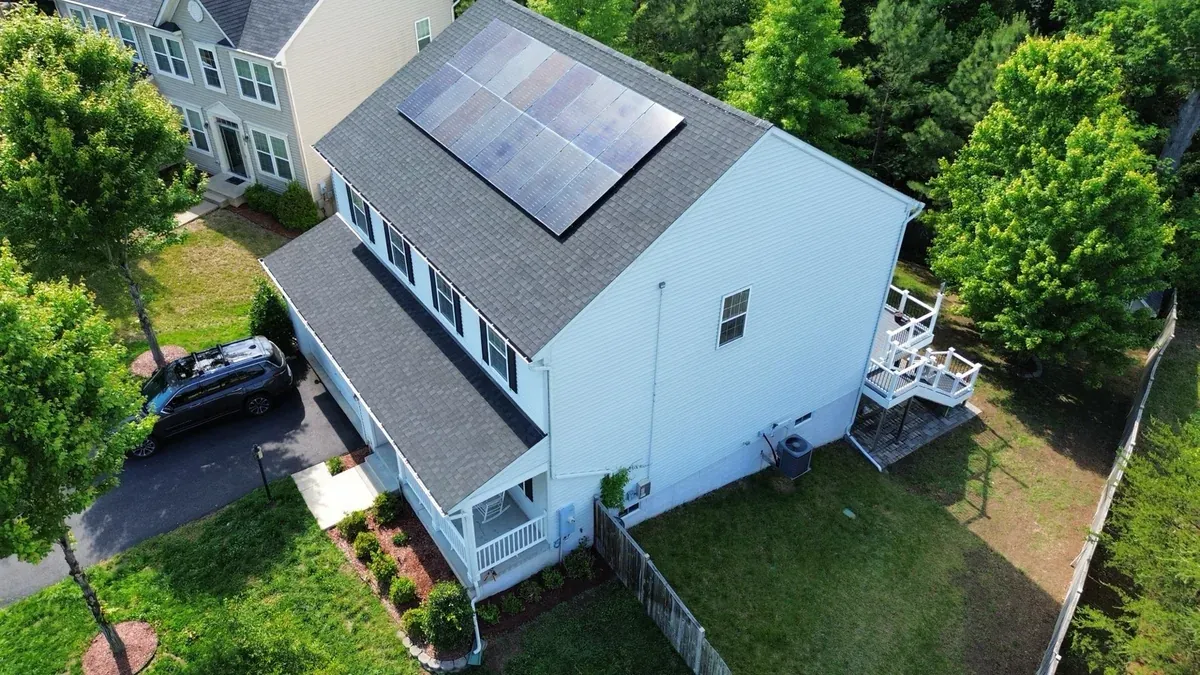 Aerial view of a white two-story home with a finished grey architectural shingle roof and a solar panel array on the main slope, front porch and rear deck below