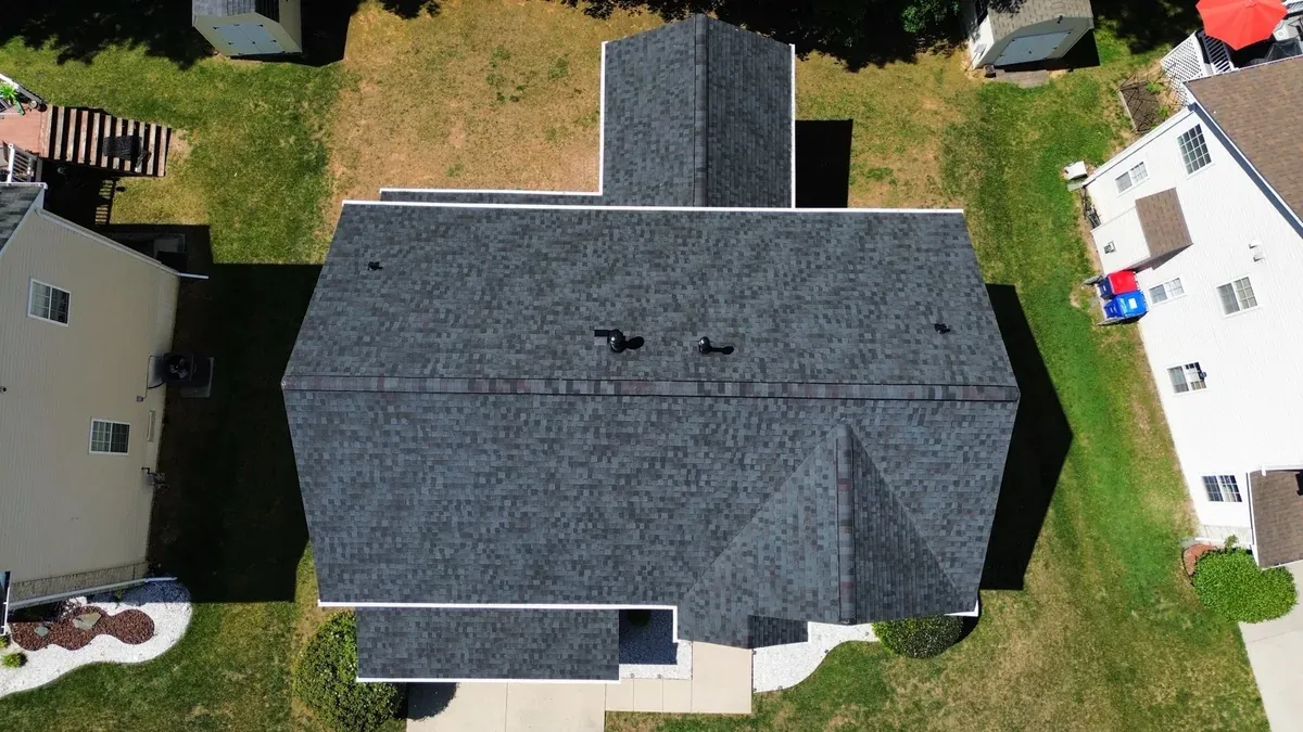 Overhead aerial view straight down onto a large finished grey architectural shingle roof with multiple planes and valleys, vent stacks and pipe boots visible, neighboring homes to either side