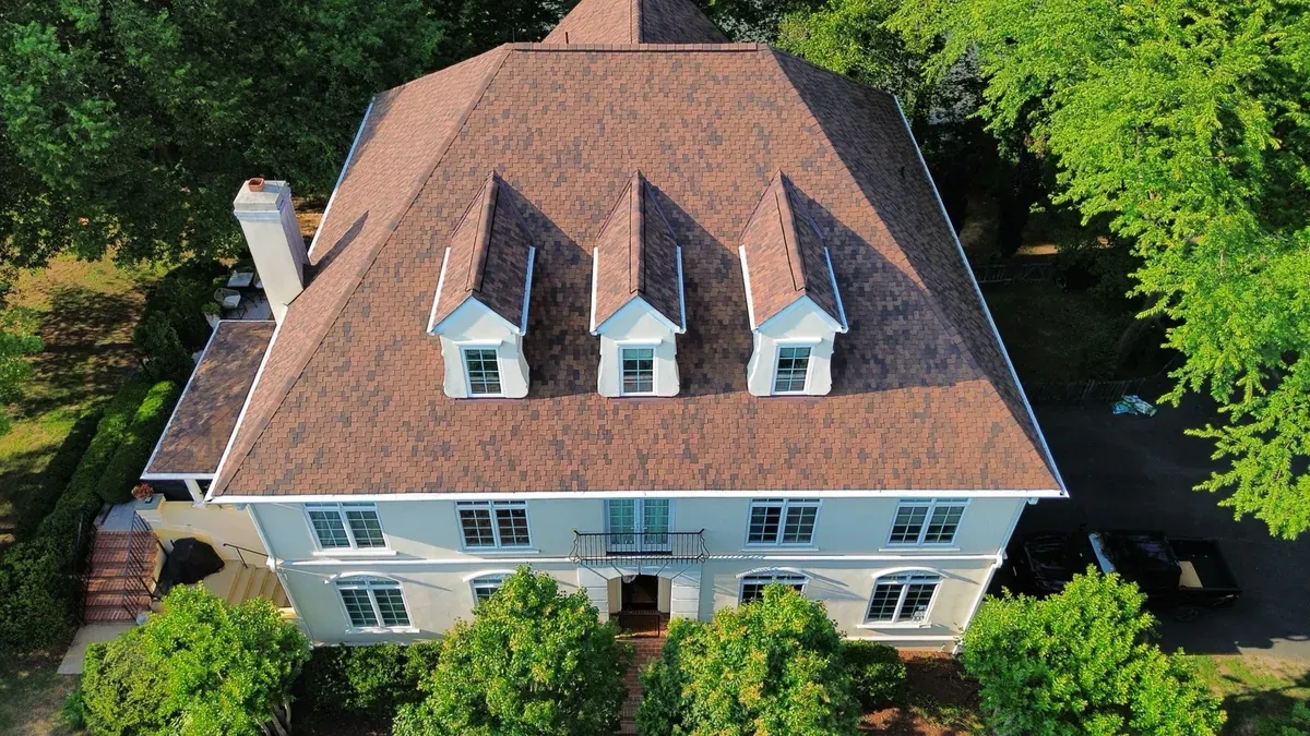 Aerial view of a large stucco home with a brown architectural shingle hip roof, three front dormers, and a masonry chimney, framed by mature trees