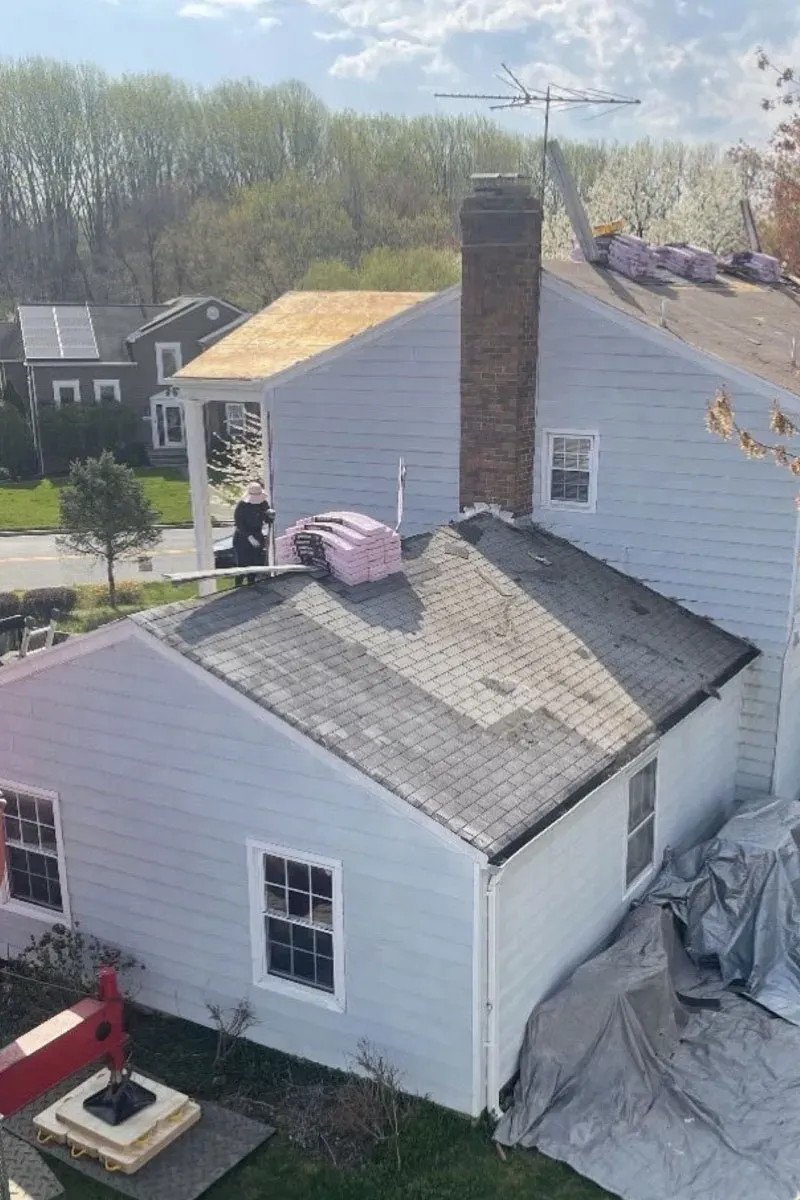 Elevated view of a white-sided home mid-tear-off, weathered grey shingles stripped back to fresh decking beside a brick chimney, with shingle bundles staged and tarps over the shrubs below