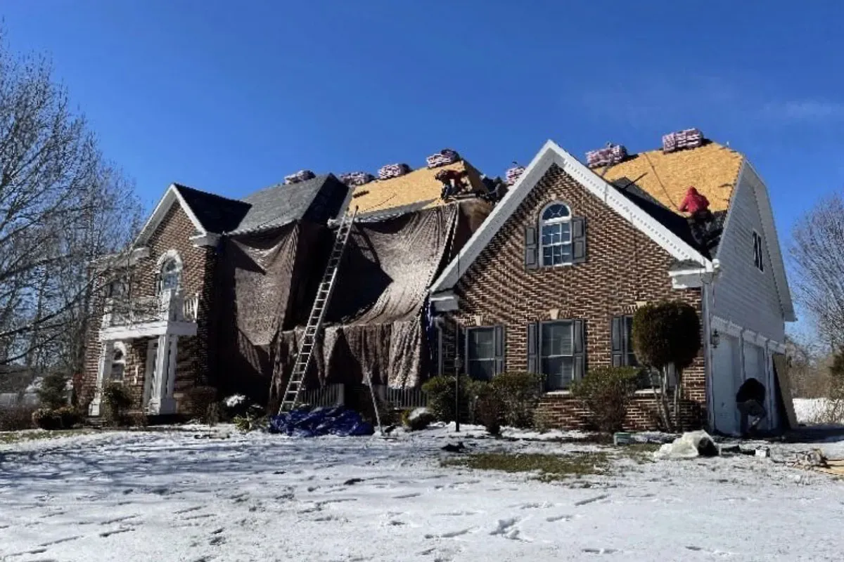Brick colonial stripped to roof decking in winter, with tarps hung down the front to protect the siding and landscaping, snow on the ground, and shingle bundles staged on the roof