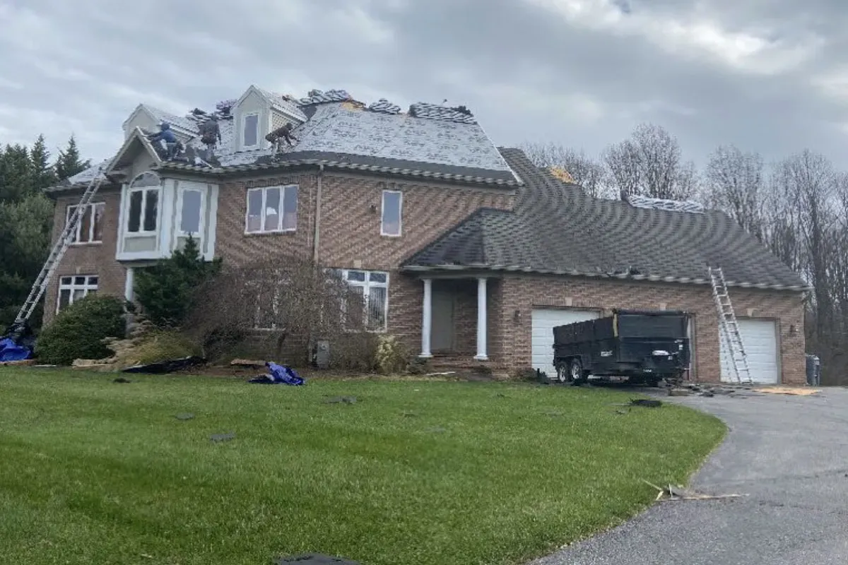 Tear-off underway on a large brick home under an overcast sky, with a dump trailer staged at the garage and ladders set at both ends of the roof
