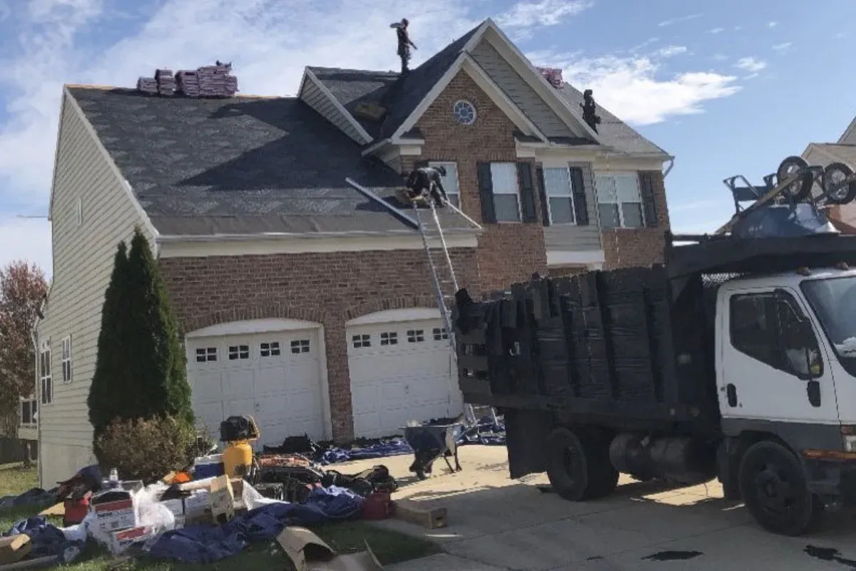 New architectural shingles being installed on a brick home, with a dump truck in the driveway collecting tear-off debris and a crew member on the ladder