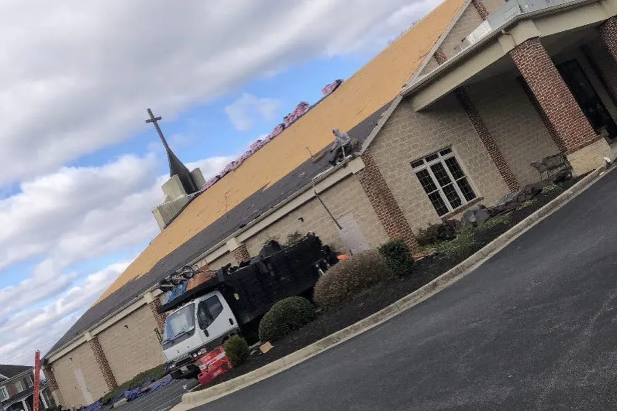 Roof deck exposed across the long slope of a brick church with a steeple and cross, a dump truck staged in the parking lot below