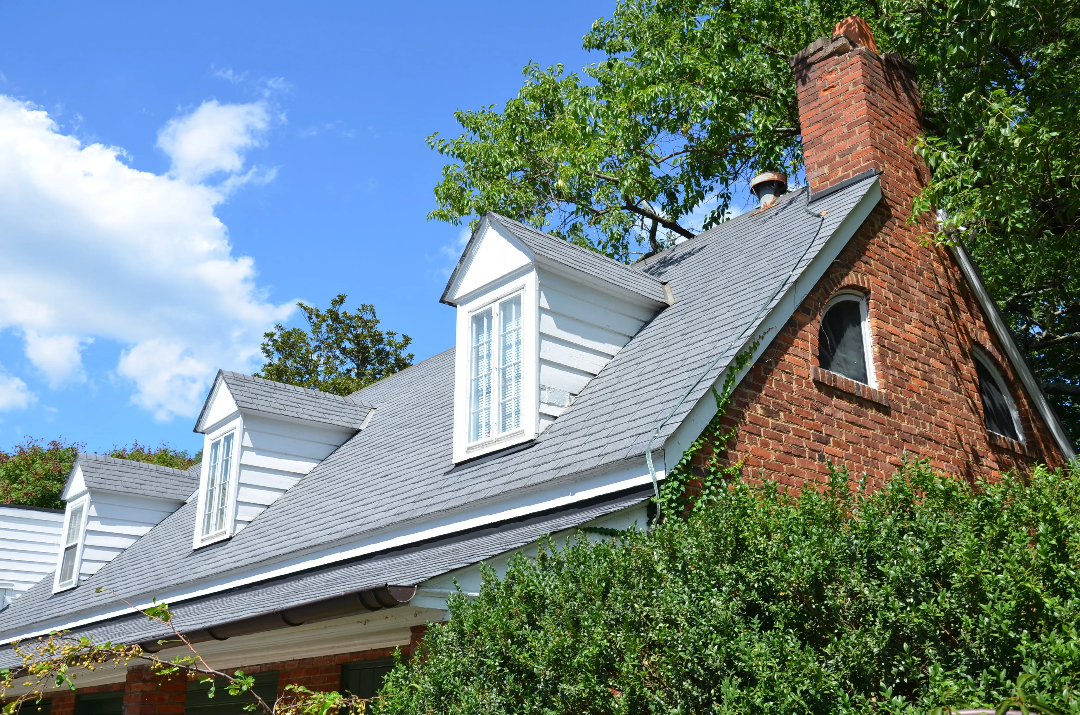 Brick Cape Cod home with a gray shingle roof, three white dormers, and a brick chimney under a blue sky