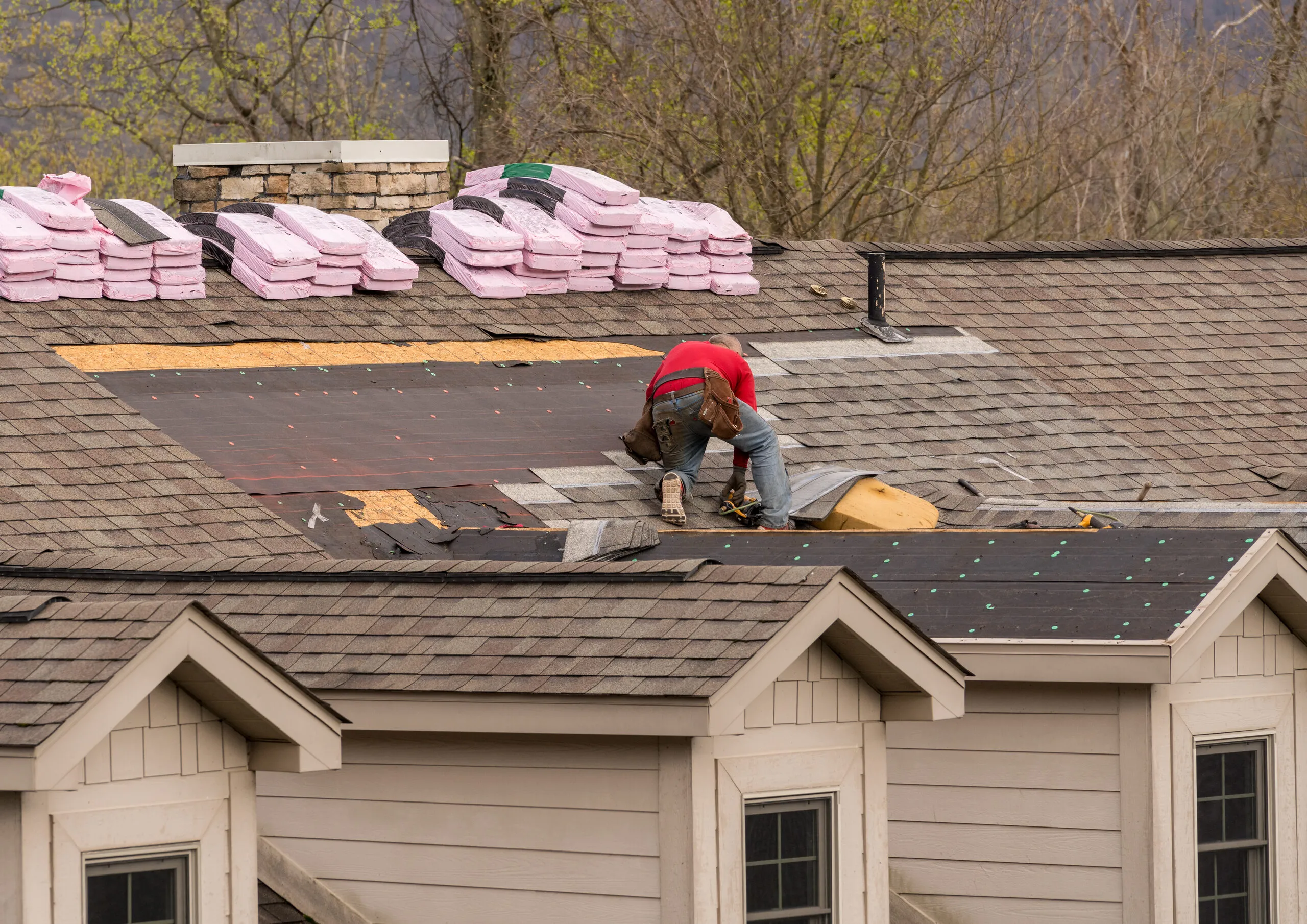 Roofer working on a shingle roof with bundles of new shingles staged along the ridge